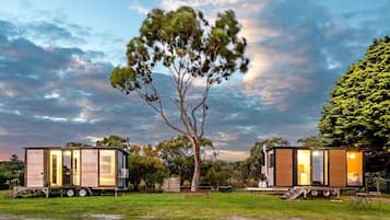 Deux maisons mobiles modernes, dotées de grandes fenêtres, sont installées sur une pelouse verdoyante, agrémentée d’arbres, avec un ciel nuageux en arrière-plan.