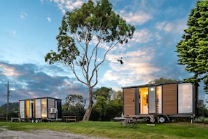 Two modern mobile homes with large windows, one with an open door, set on a trailer in a grassy area with trees and a clear sky.