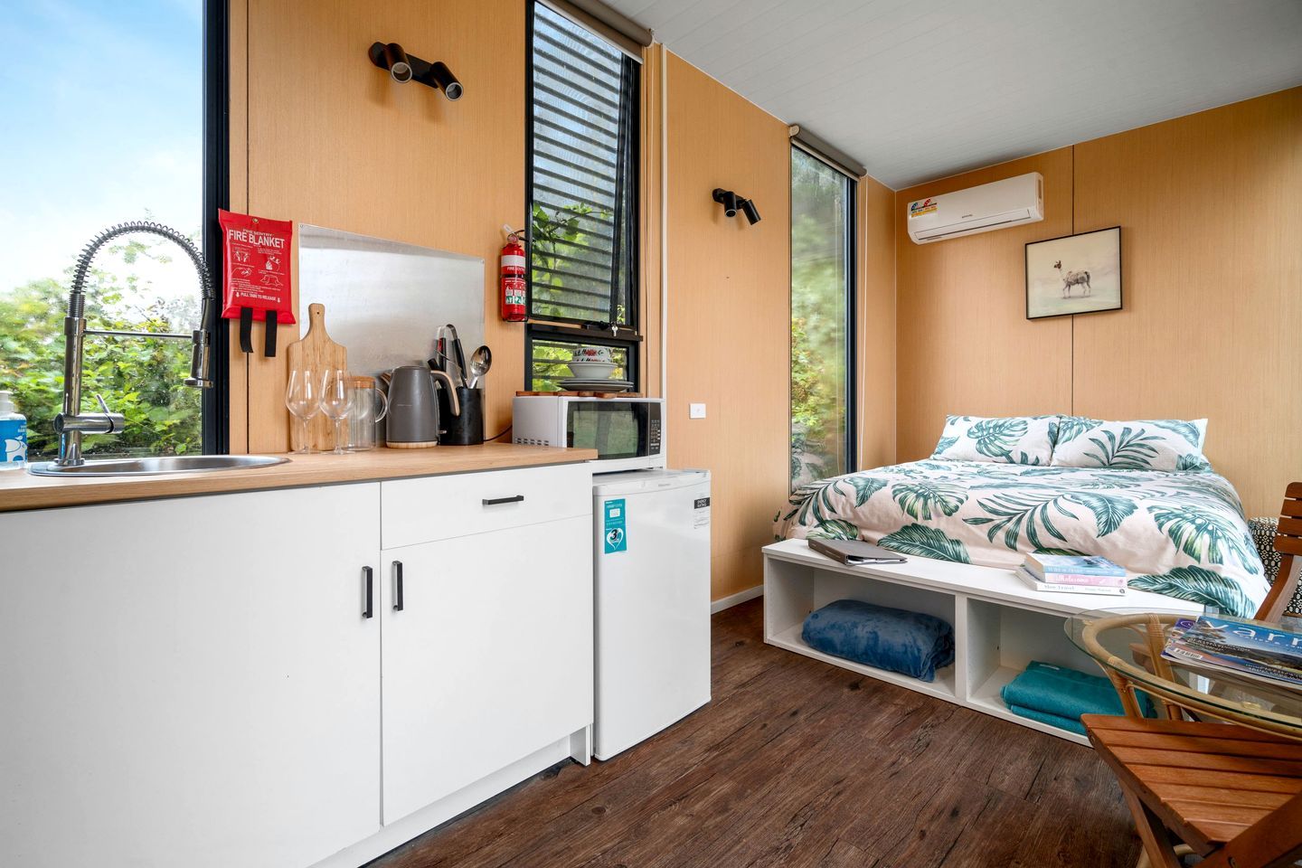 A compact room with a kitchenette, a bed with a tropical print, a window with blinds, and a mounted fire extinguisher.