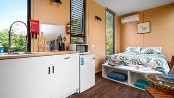 A compact room with a kitchenette, a bed with a tropical print, a window with blinds, and a mounted fire extinguisher.