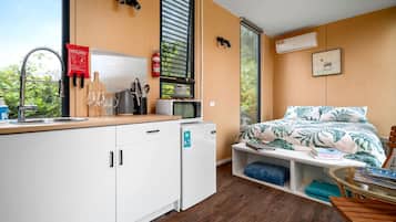 Una habitación compacta con kitchenette, una cama con estampado tropical, una ventana con persianas y un extintor de incendios montado.