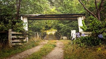 A rustic wooden bridge with a white archway, surrounded by lush greenery and a gravel path leading through a wooded area.