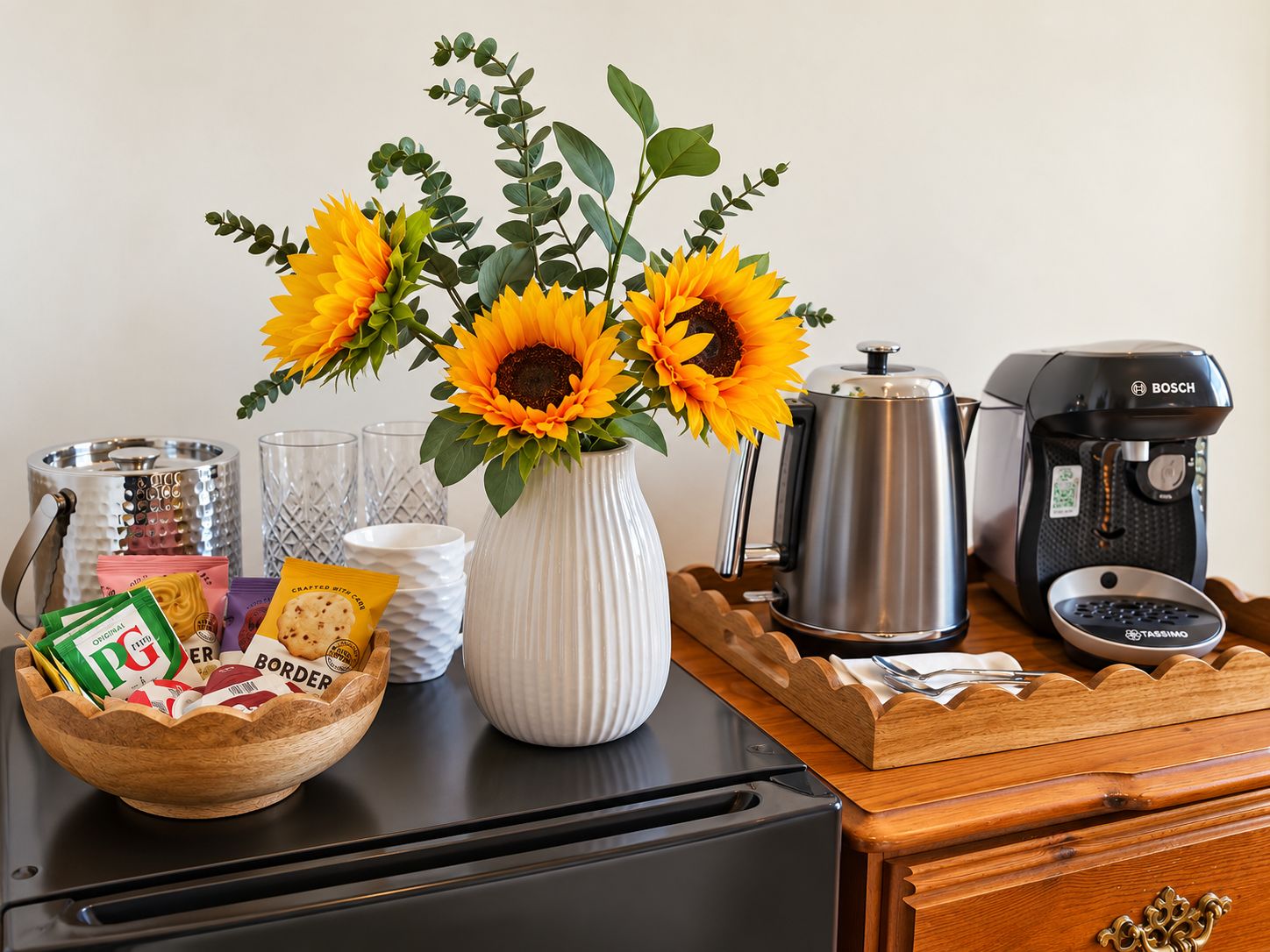 Una encimera de cocina con una cafetera, una olla, un cuenco de aperitivos, un florero con girasoles y un molinillo de café.