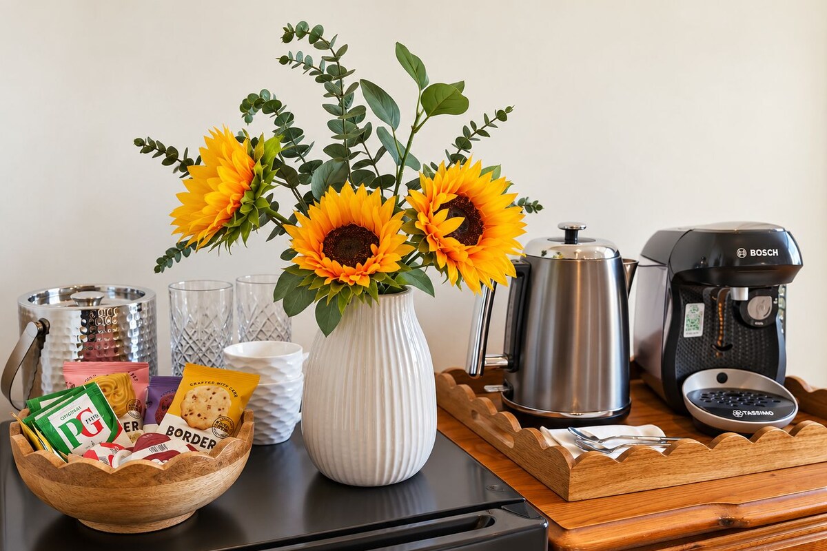 A kitchen counter with a coffee maker, a pot, a bowl of snacks, a vase with sunflowers, and a coffee grinder.