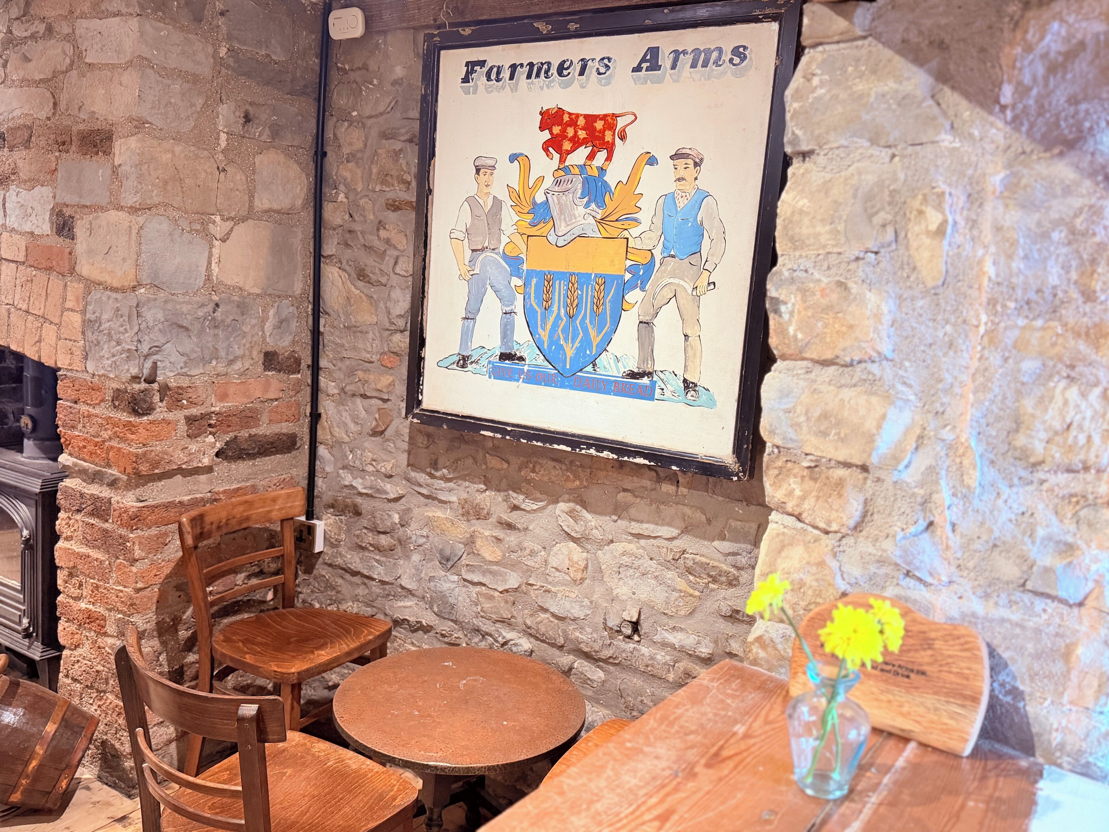 A rustic interior with stone walls, wooden furniture, and a framed poster of 'Farmers Arms' featuring a coat of arms.
