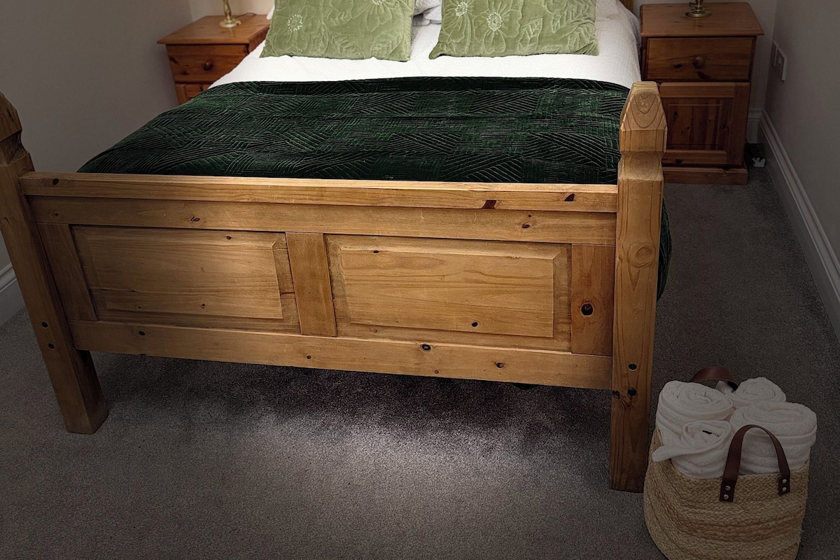 A wooden bed with green bedding, two bedside tables, and a small woven bag on the floor.