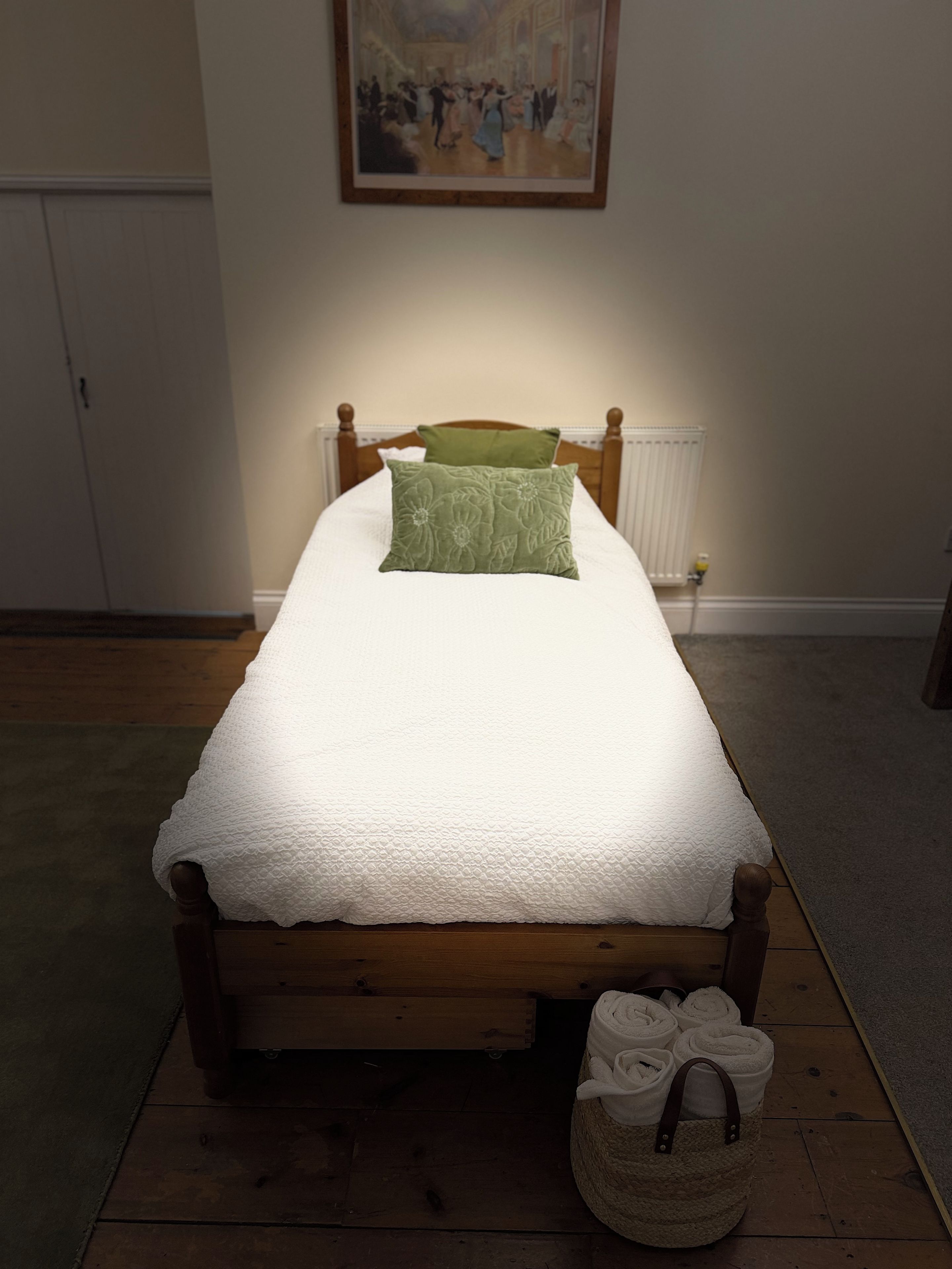 A single bed with a wooden frame, a white quilt, and a green pillow. A radiator is visible on the wall behind the bed.