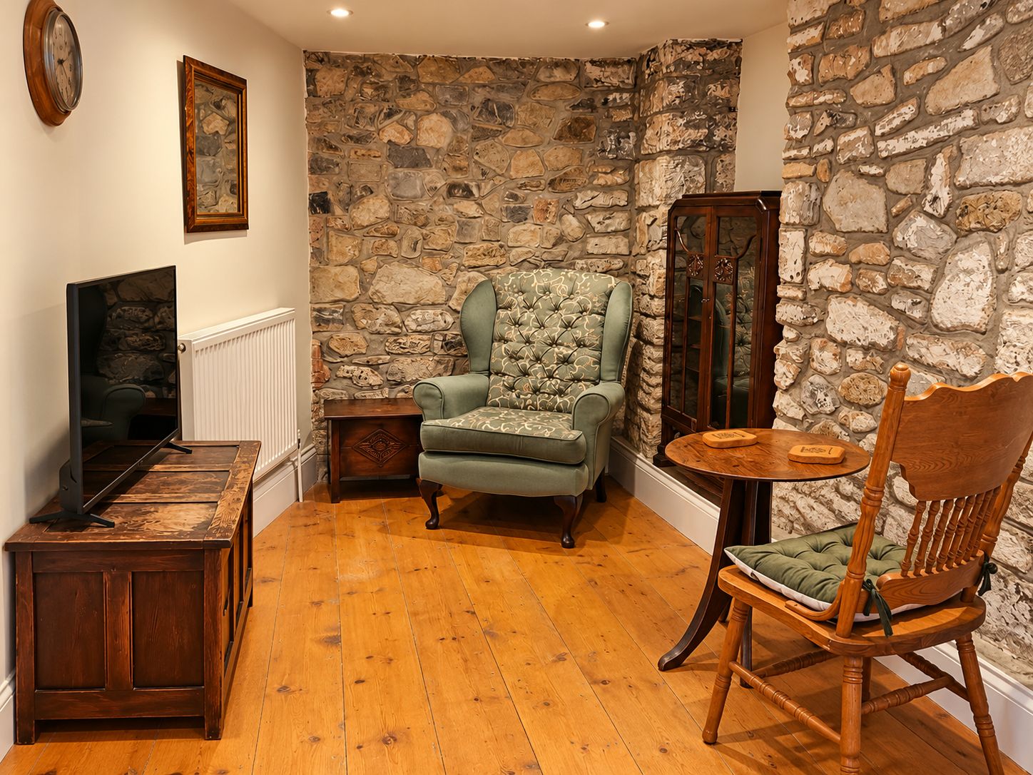 A room with a stone wall, a green upholstered armchair, a wooden dining table with a green cushion, a wooden chair, a wooden side table, and a television on a wooden stand.
