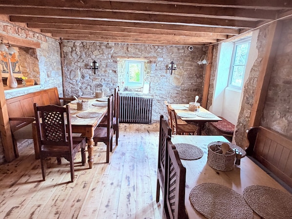 A rustic dining area with wooden tables and chairs, stone walls, and a window providing natural light.