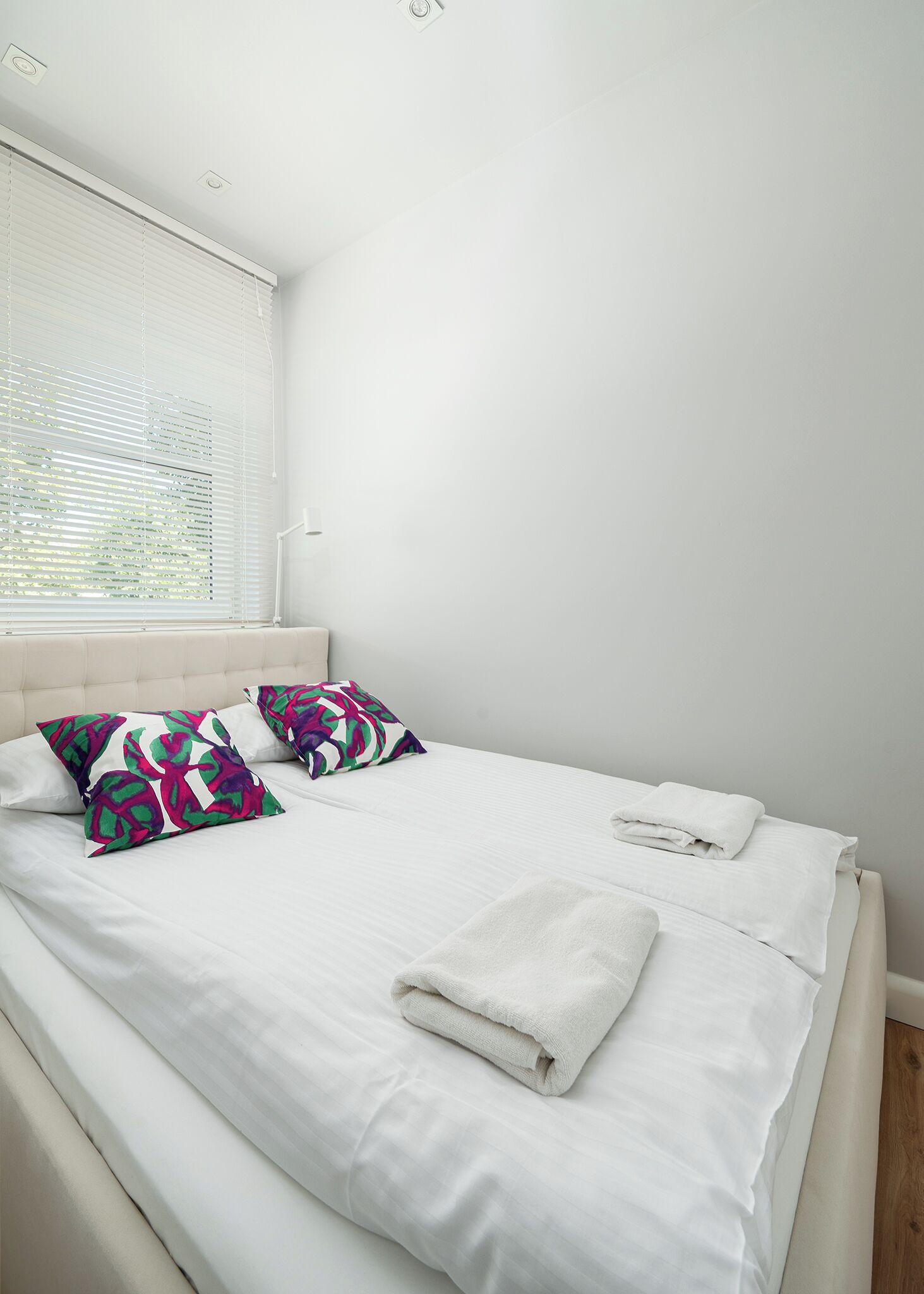 Een netjes opgemaakt bed met witte lakens en twee kussens, een raam met zonwering en een lichtschakelaar aan de muur.