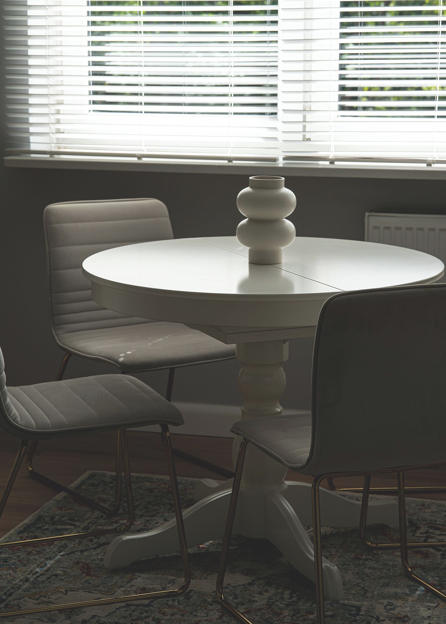 Uma mesa de jantar redonda com base branca em pedestal, rodeada por quatro cadeiras com pés de metal.