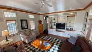 A living room with a ceiling fan, a television on a cabinet, a checkered rug, and a brown sofa. - Peak and Pond Mountain View Retreat hot tub theater sauna. (Killington)