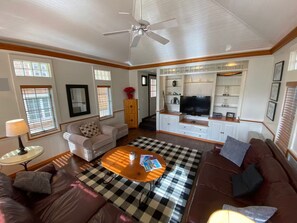 A living room with a ceiling fan, a television on a cabinet, a checkered rug, and a brown sofa. - Peak and Pond Mountain View Retreat hot tub theater sauna. (Killington)