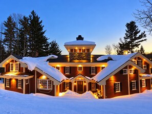 Uma grande casa de madeira de vários andares, com telhados cobertos de neve e uma torre central, rodeada por uma paisagem nevada e árvores.