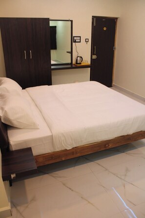 A bedroom with a wooden bed frame, a white mattress, a wooden wardrobe, and a small shelf with a kettle and cups.