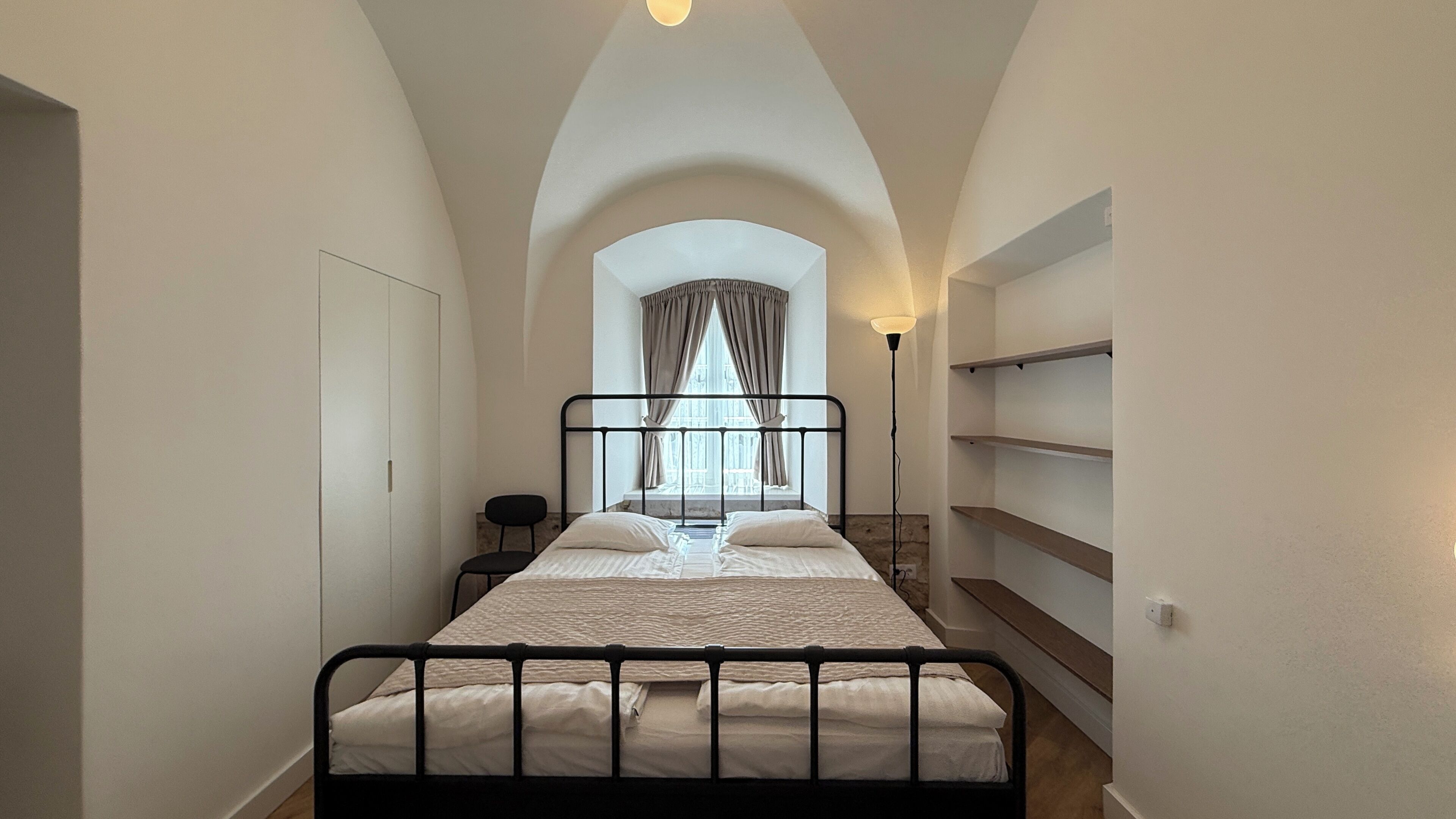Um quarto pequeno com uma cama, uma cadeira, uma escrivaninha e uma janela com cortinas.