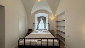 A narrow room with a bed, a chair, a desk, and a window with curtains.