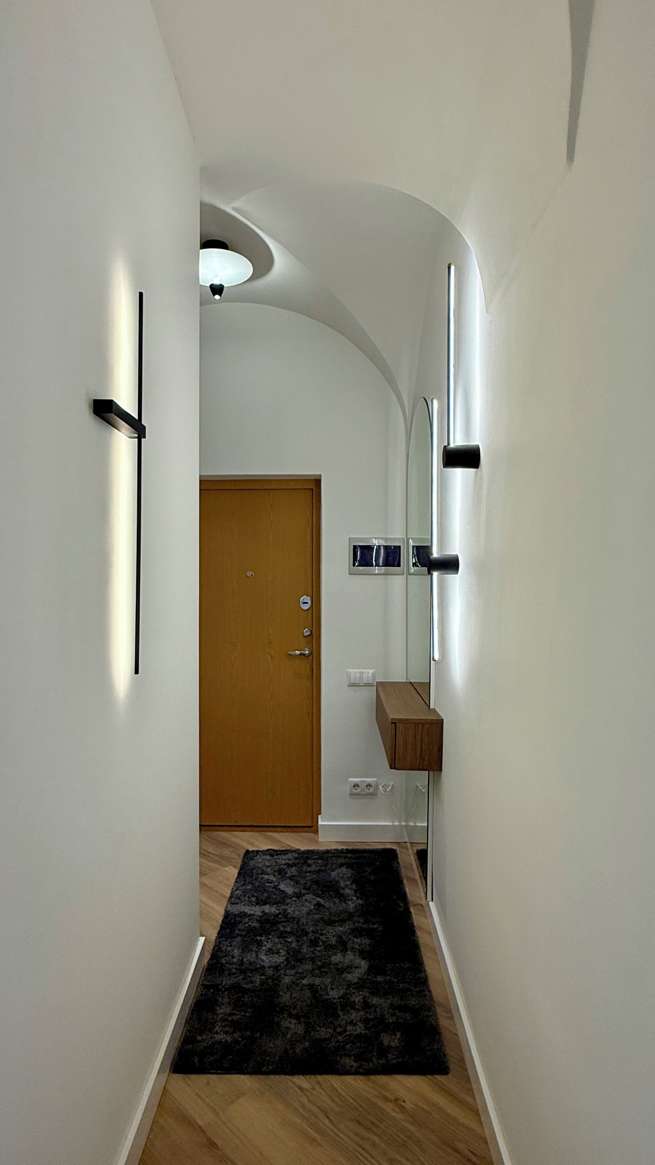 Un couloir avec une porte en bois, un tapis noir et une étagère en bois sur laquelle est posé un miroir.