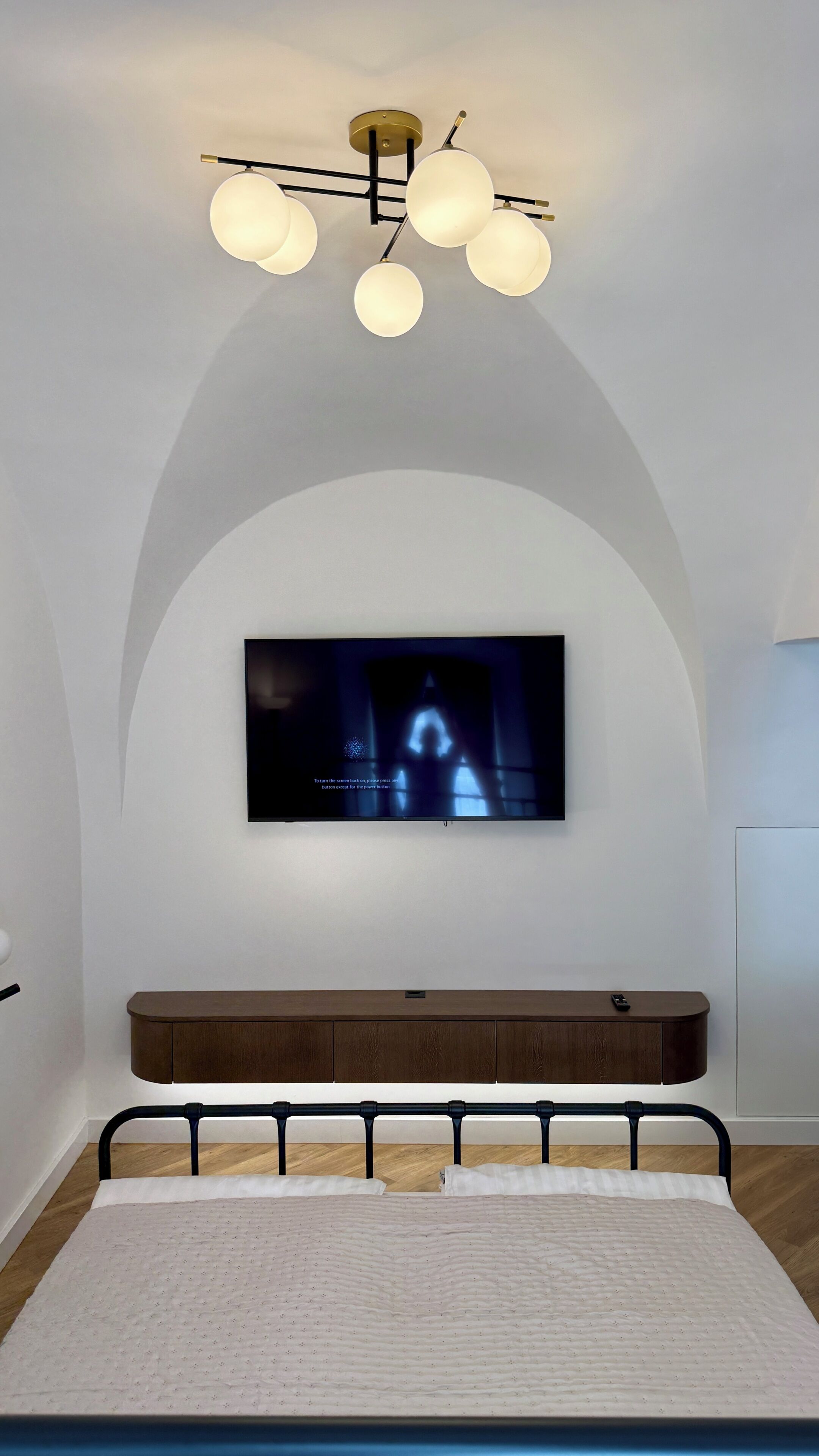 Un dormitorio con una televisión montada en una pared abovedada, una cama con cabecera de madera y una lámpara de techo moderna.