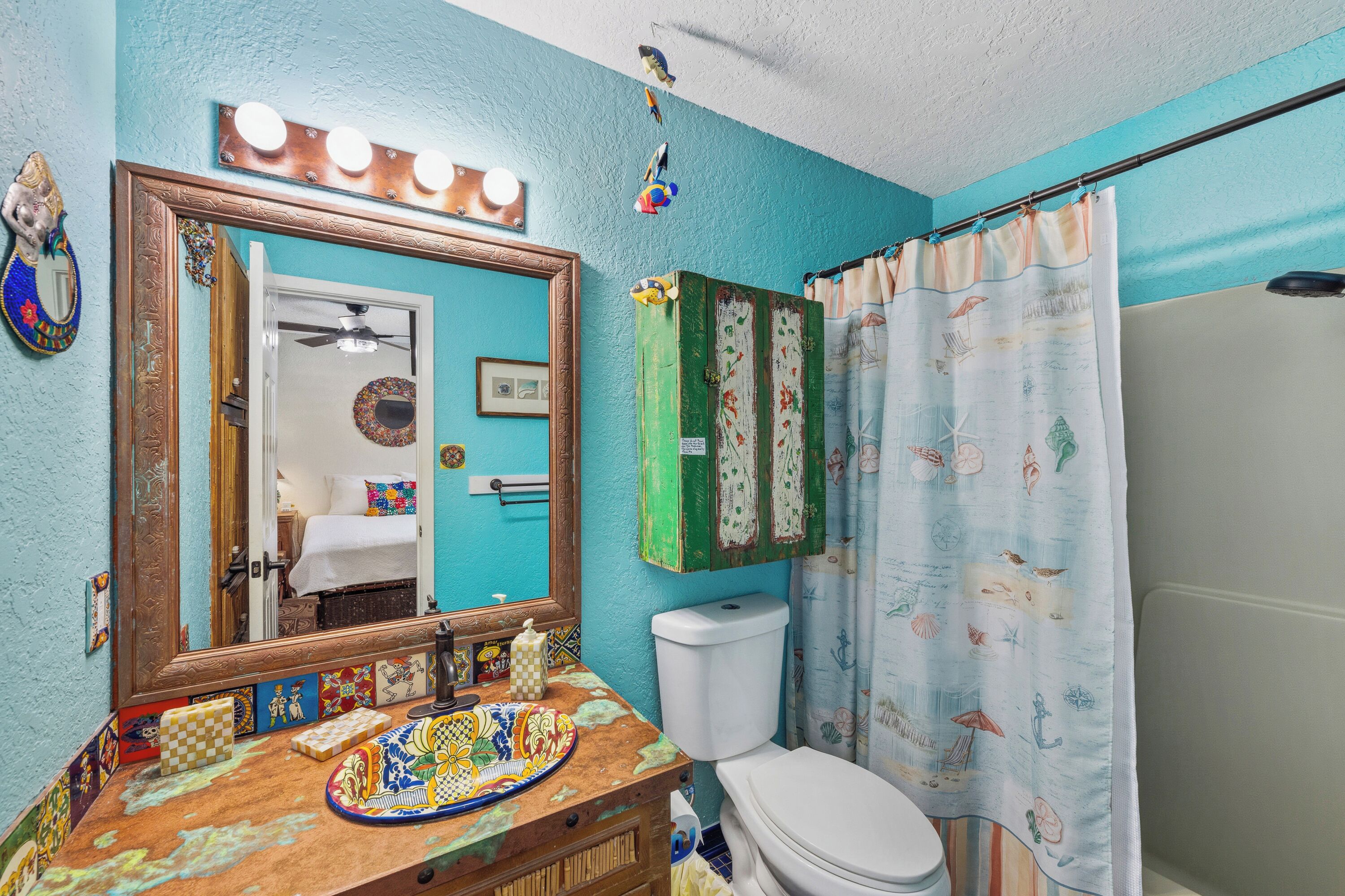 A bathroom with a turquoise wall, a wooden vanity with a colorful tile top, a white toilet, and a shower curtain with a beach theme.