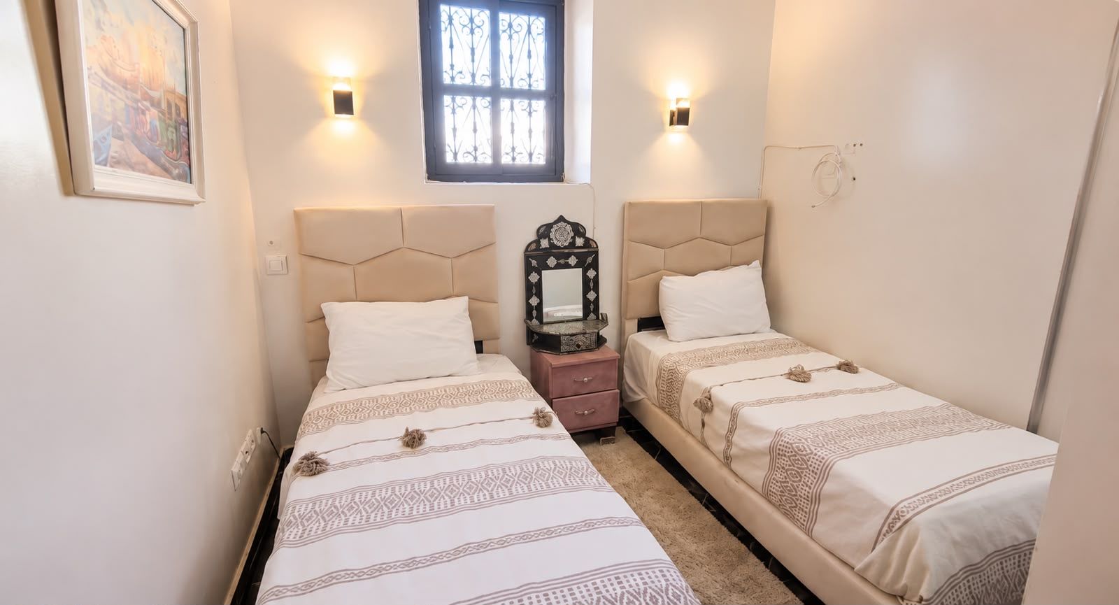 Une chambre à coucher avec deux lits, chacun doté d’une tête de lit, d’une table de chevet, d’un miroir et d’une fenêtre ornée de barreaux en fer décoratifs.