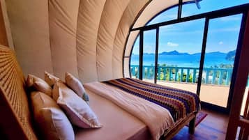 A bedroom with a large curved window offering a view of the ocean and mountains.