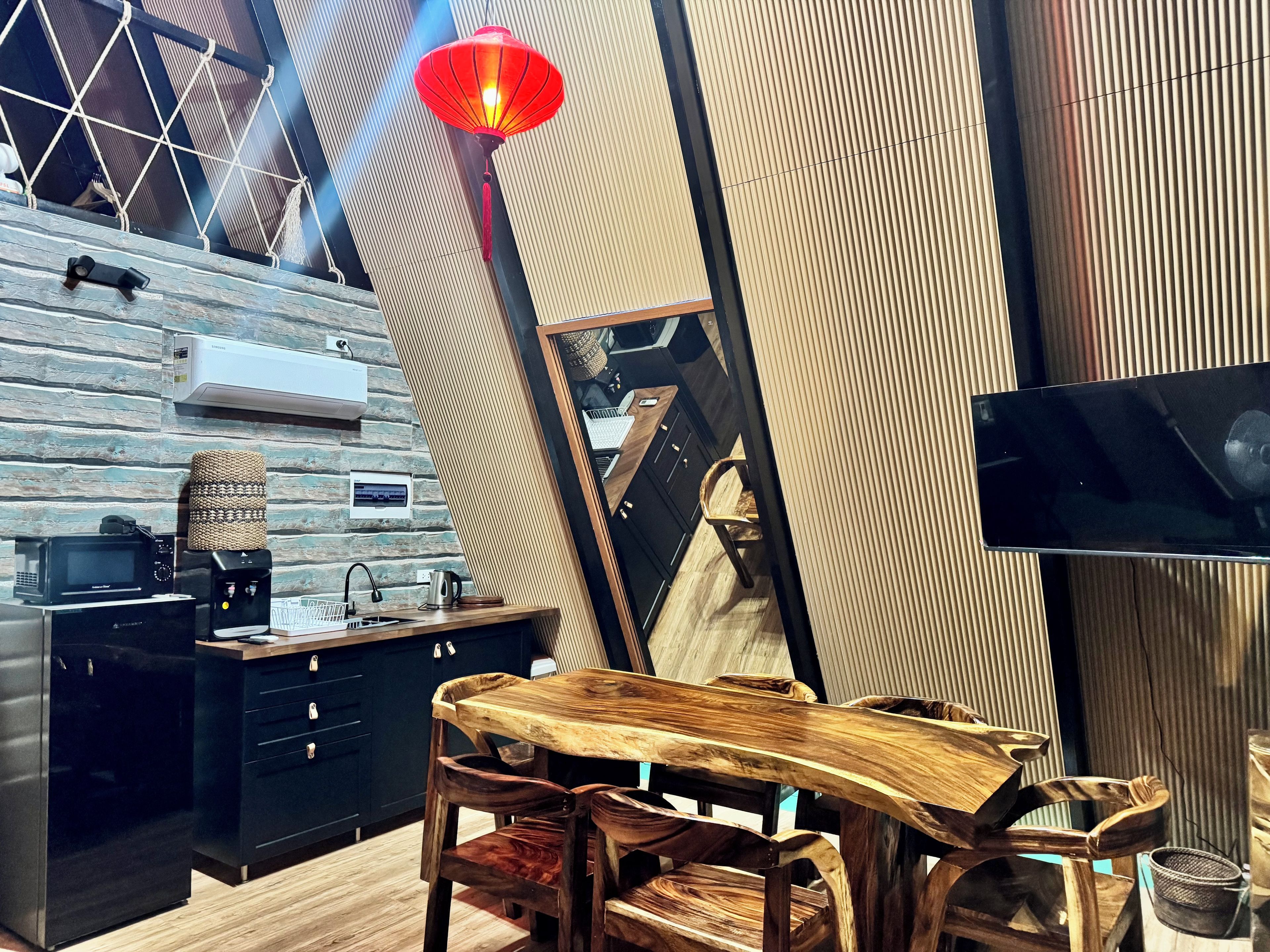 A modern kitchen with a wooden table and chairs, a TV mounted on the wall, and a red hanging light fixture.