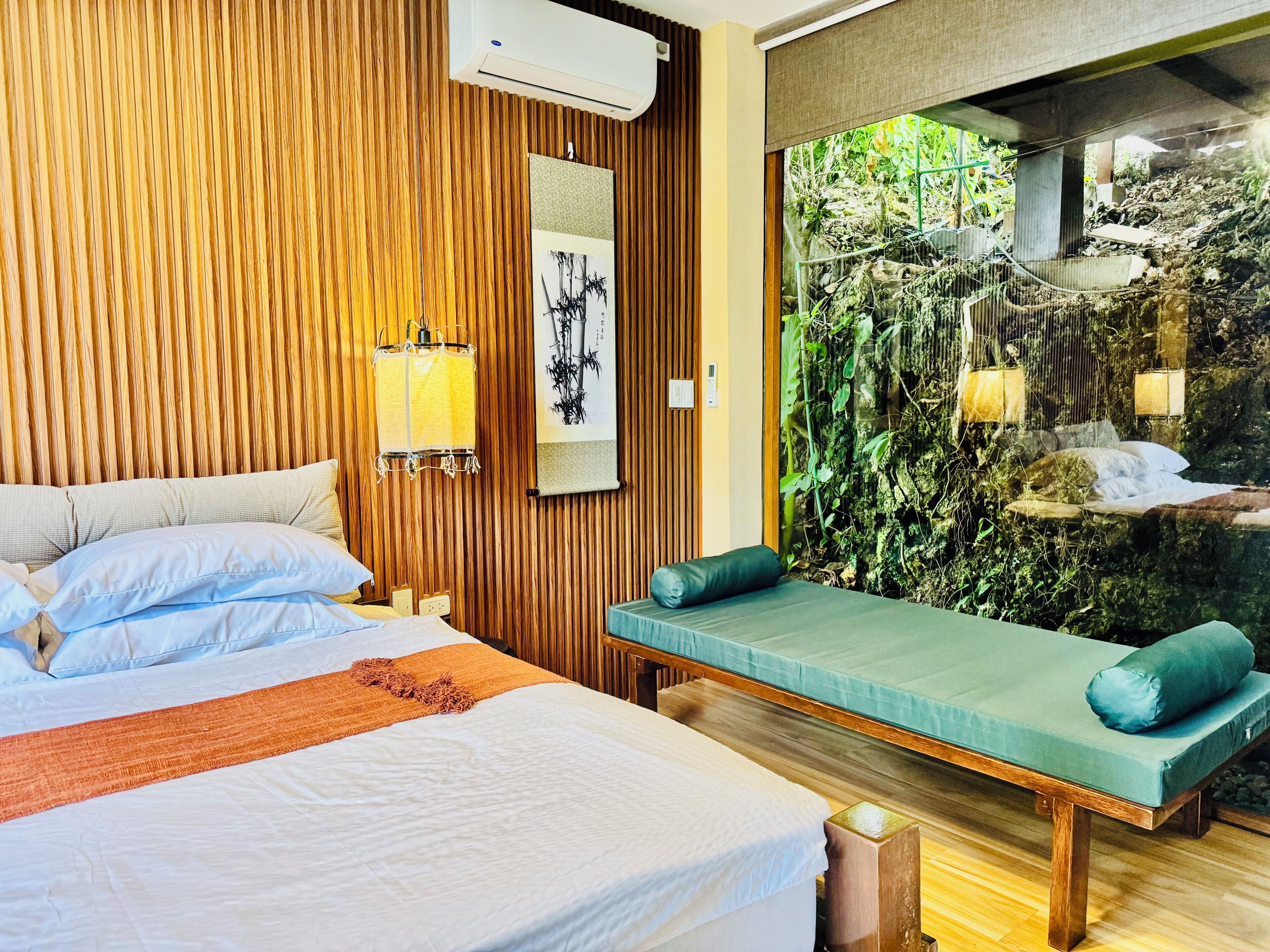 A hotel room with a bed, a bench, a wall-mounted air conditioner, and a large window with a view of a rocky garden.