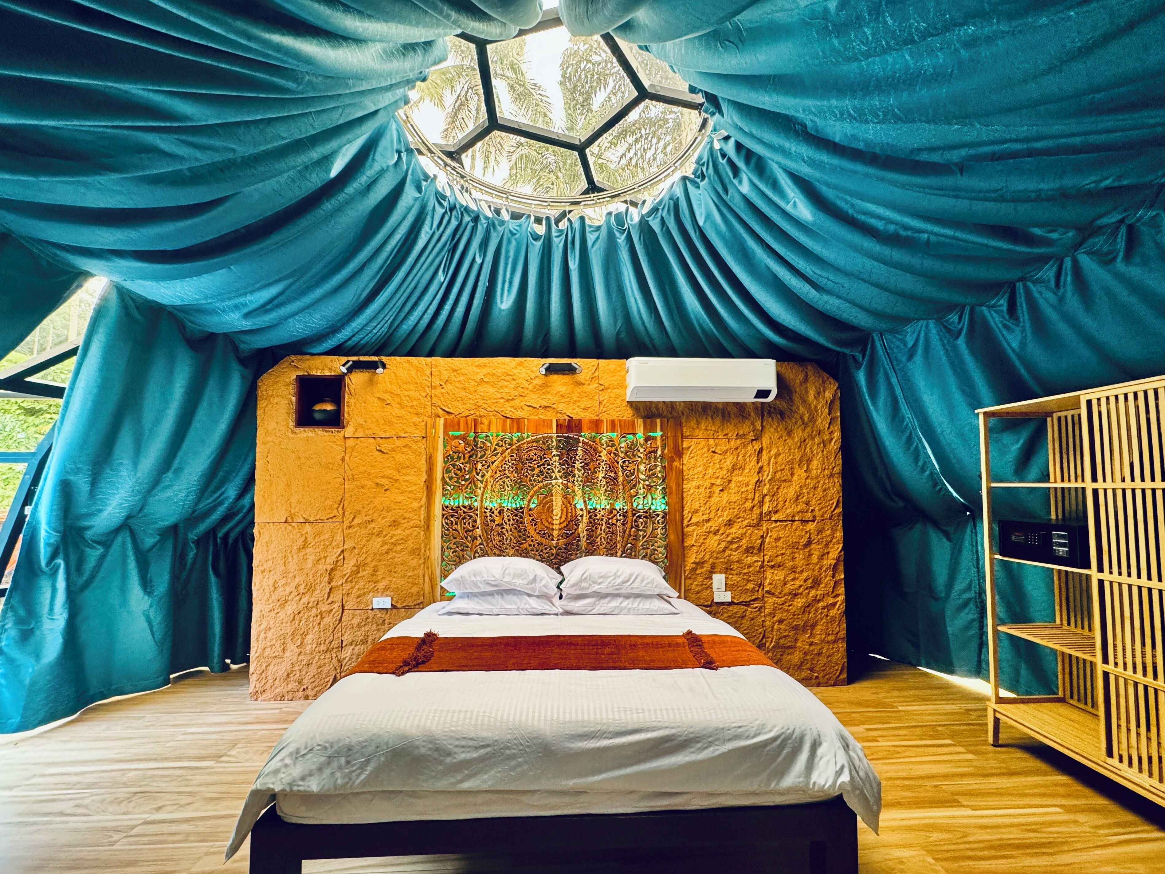 A room with a bed, a wooden floor, a wall with a hexagonal window, and a ceiling with draped fabric.