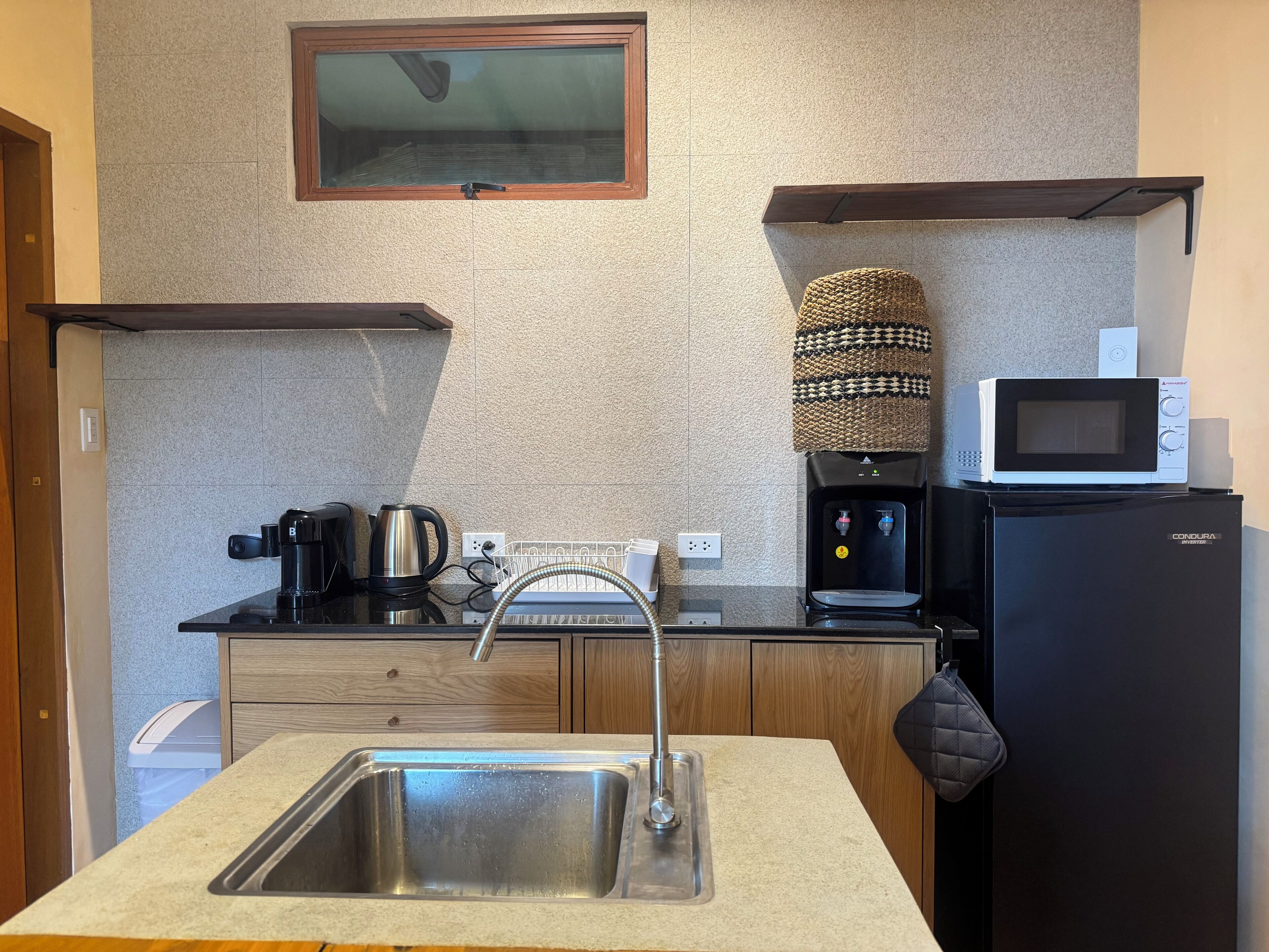A compact kitchen with a stainless steel sink, a coffee maker, a kettle, a microwave, and a water dispenser.