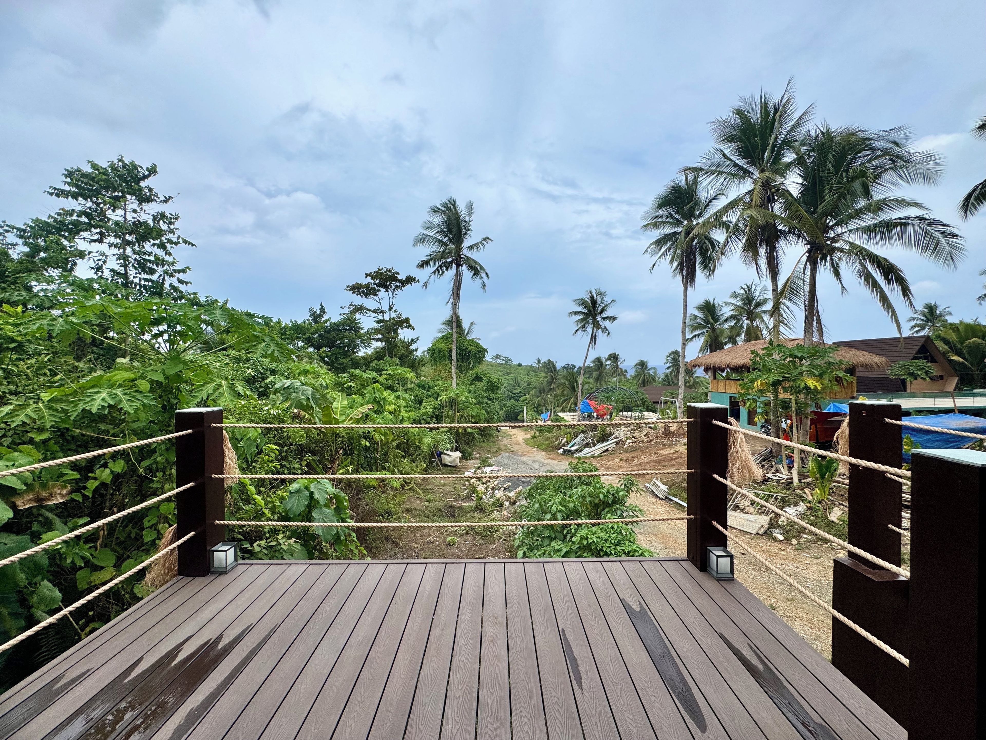 Una terraza de madera con barandas de cuerda, con vistas a un paisaje exuberante con palmeras y una estructura con techo de paja.