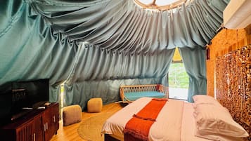 A bedroom with a bed, a wooden wardrobe, a seating area with cushions, and a window with curtains.
