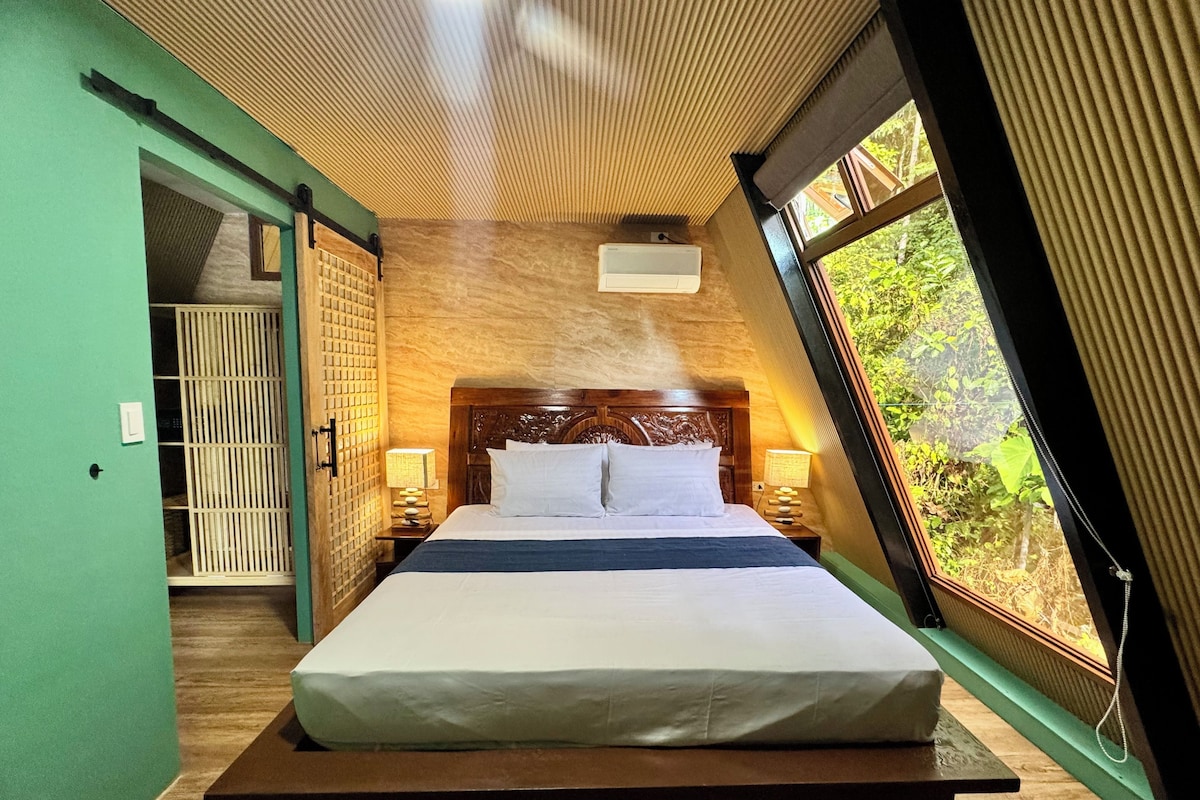 A bedroom with a large bed, wooden headboard, two bedside lamps, a window with a view of greenery, and a ceiling fan.