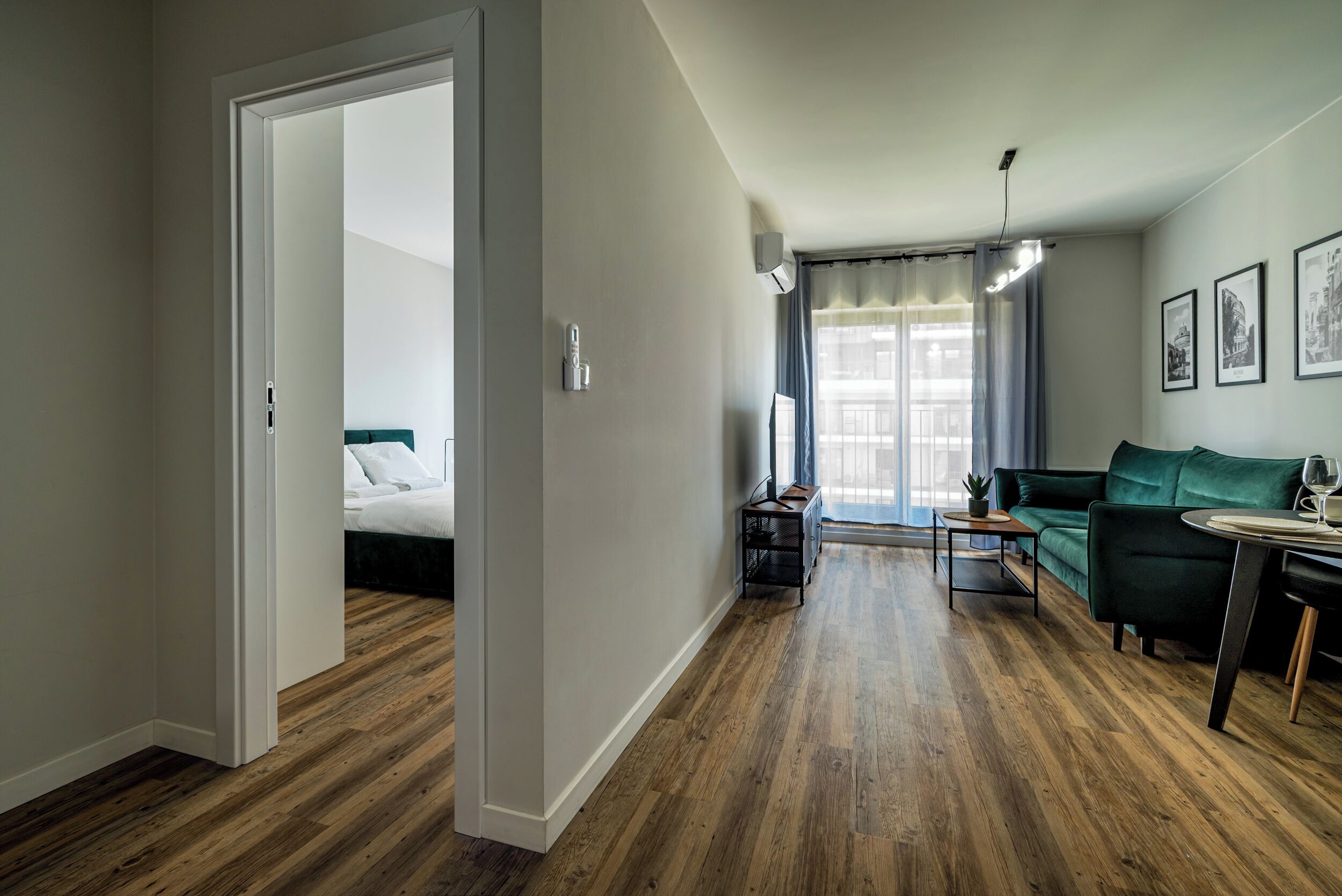 Una sala moderna con piso de madera, un sofá verde, una mesita y una cama en la habitación contigua.
