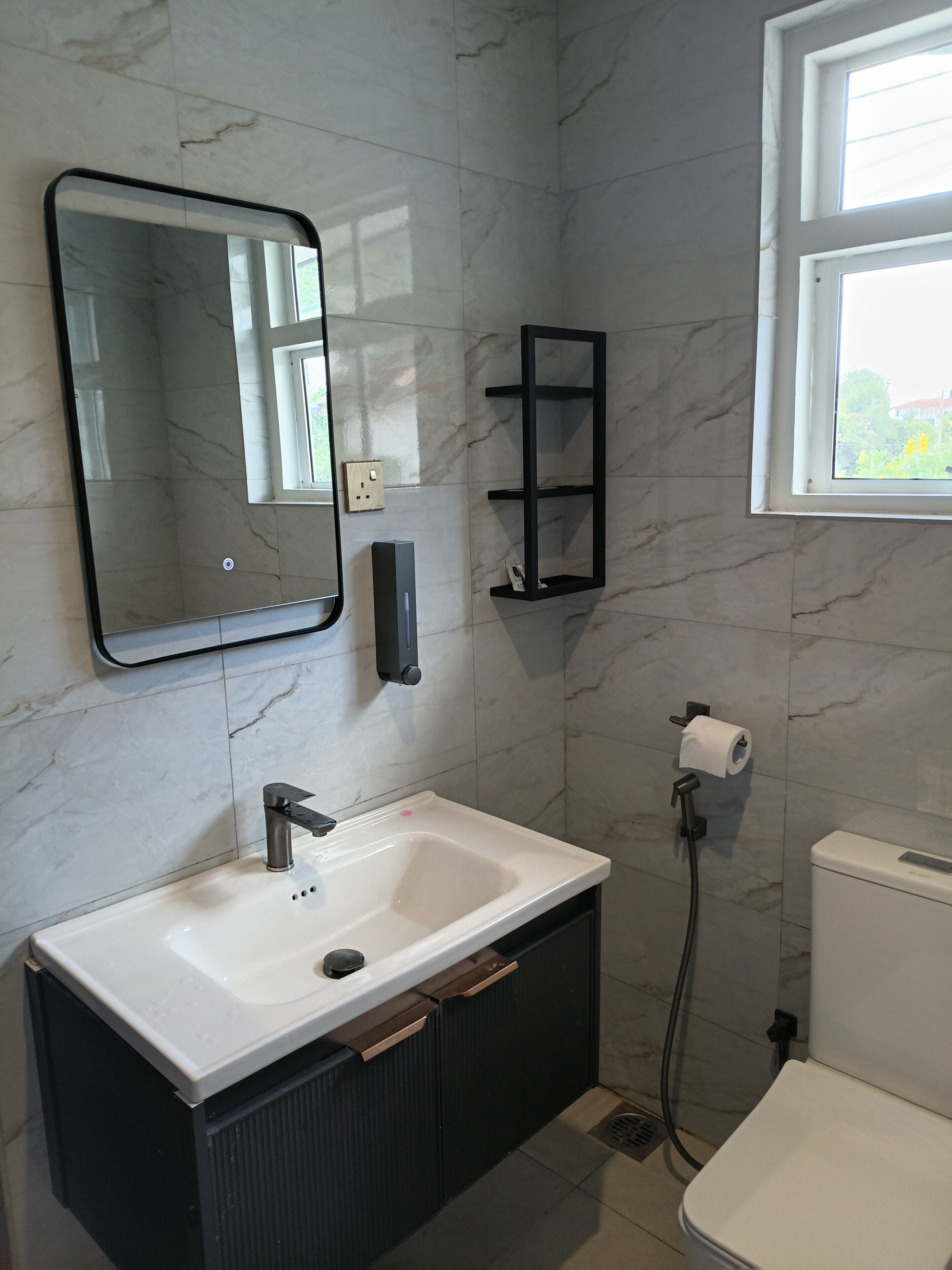 A bathroom with a white sink, a mirror, a black medicine cabinet, a toilet, and a window.