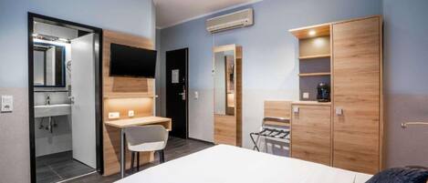 Habitación de hotel con cama, escritorio, silla, armario, espejo y baño visible a través de una puerta abierta.