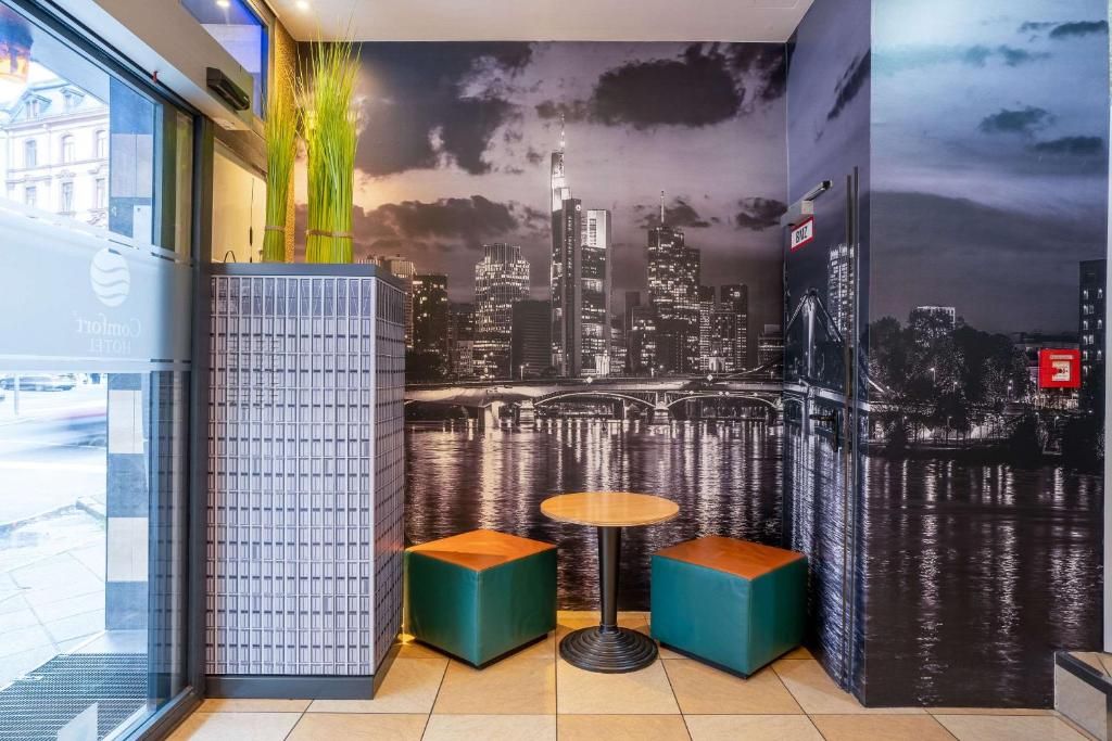 Un interior moderno con un mural de la ciudad, dos zonas de estar con cojines verdes y una pequeña mesa redonda.