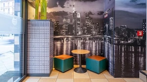 Un interior moderno con un mural de la ciudad, dos zonas de estar con cojines verdes y una pequeña mesa redonda.