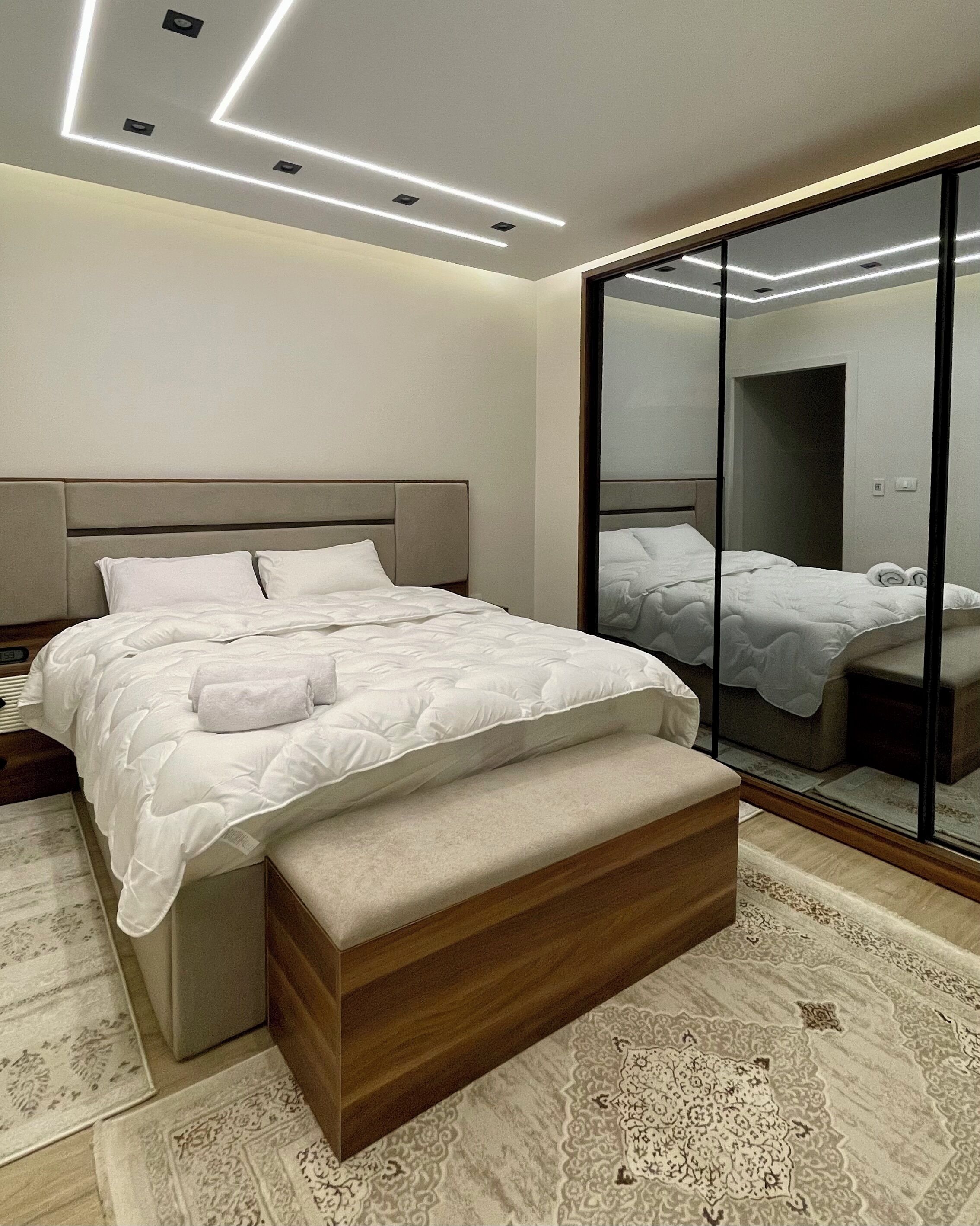 A modern bedroom with a large bed, a wooden bench, a mirror, and a patterned rug.