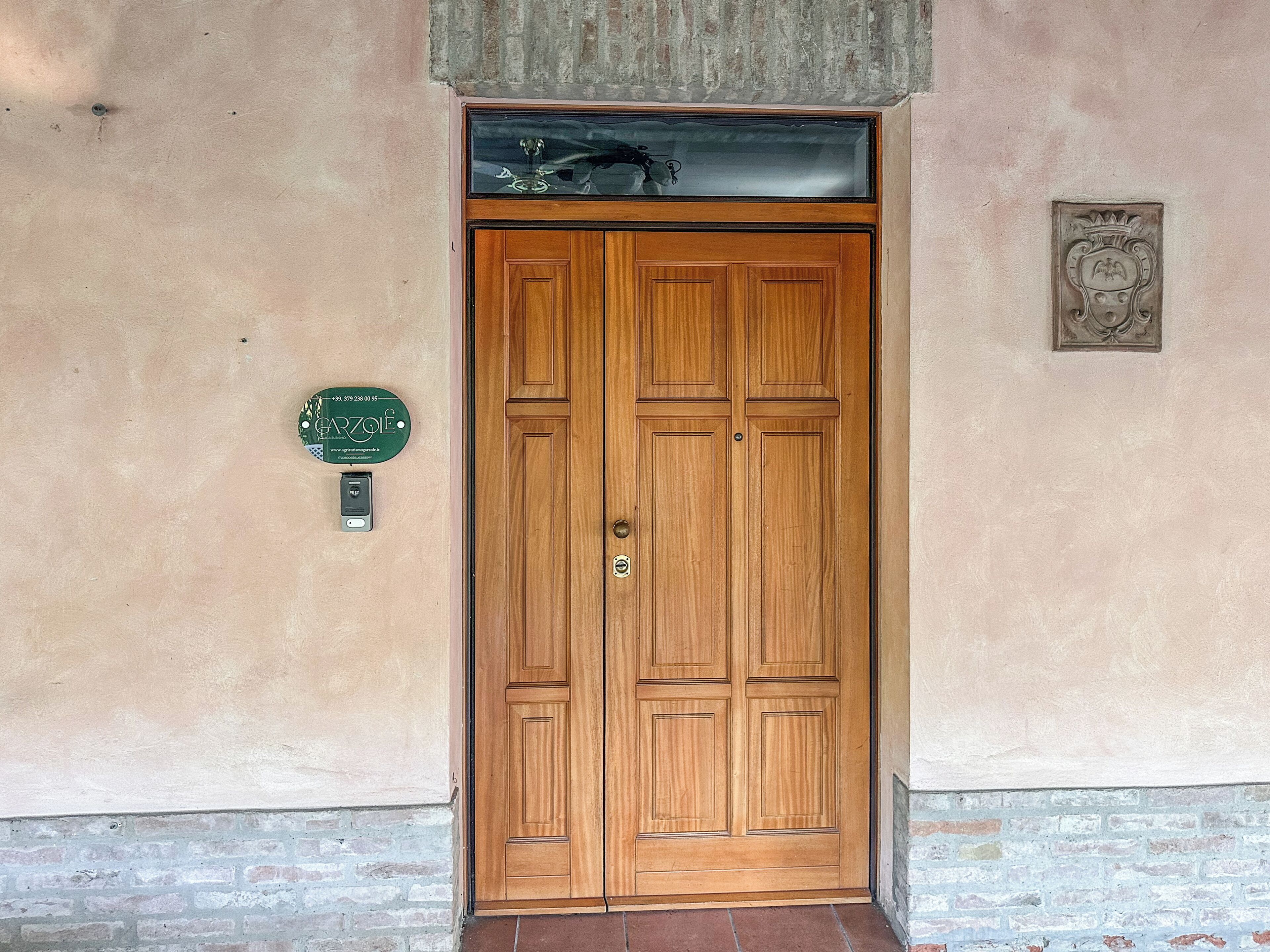 A wooden double door with a keypad lock, a green plaque with contact information, and a decorative metal plate above the door.