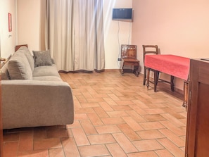 Una sala de estar con un sofá, una mesa de comedor con mantel rojo, un televisor montado en la pared y una silla de madera.