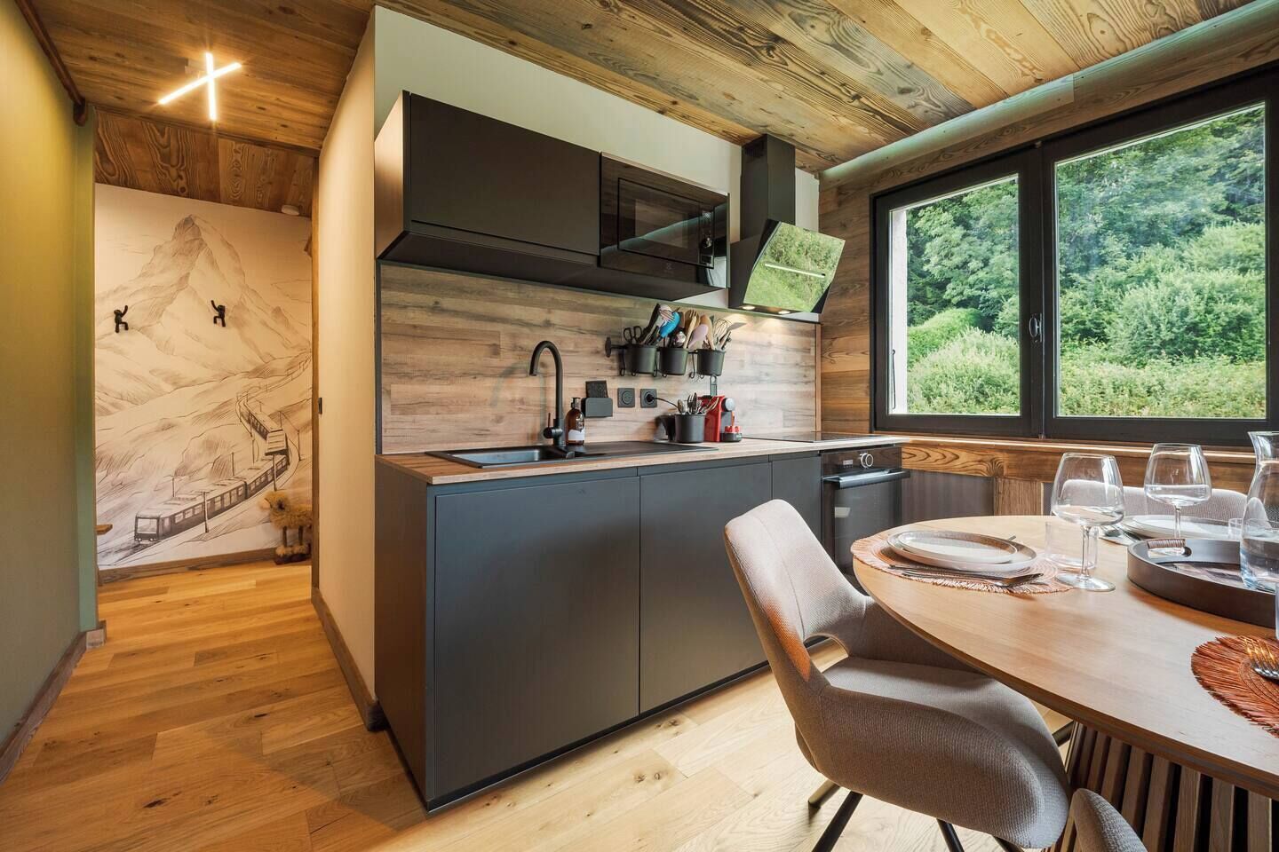 Una cucina moderna con isola centrale, zona pranzo con tavolo rotondo e vista su spazi verdi attraverso ampie finestre.