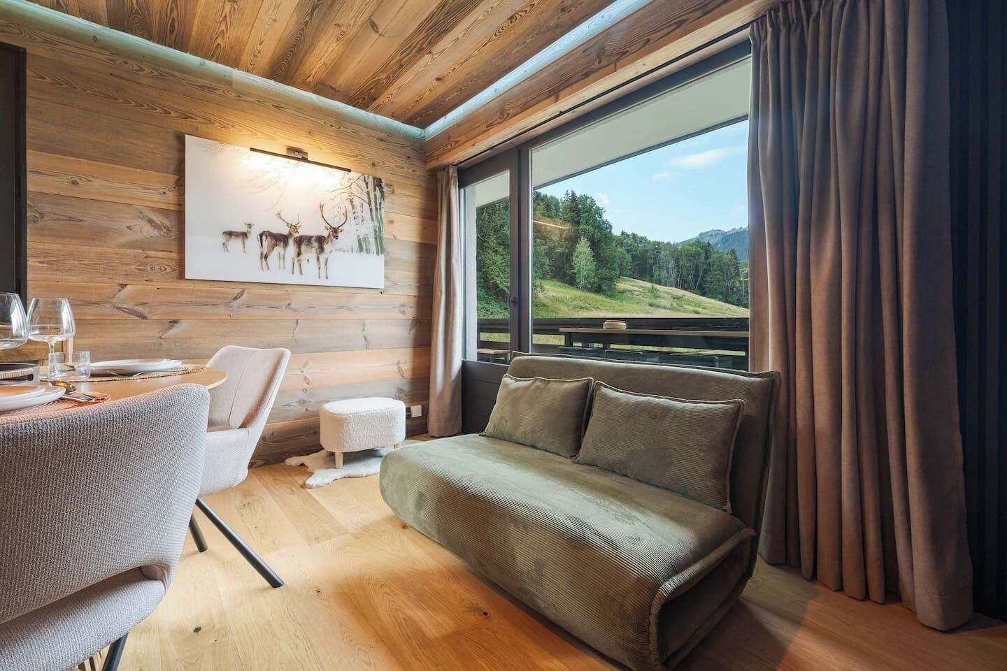Een gezellige kamer met een houten plafond, een bank, een eettafel met stoelen en een schilderij van herten aan de muur.