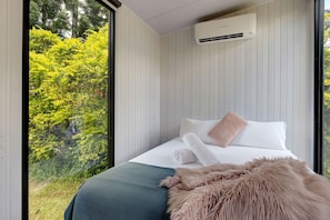 A bedroom with a large window, a bed with white bedding, a fluffy pillow, and an air conditioning unit on the wall.
