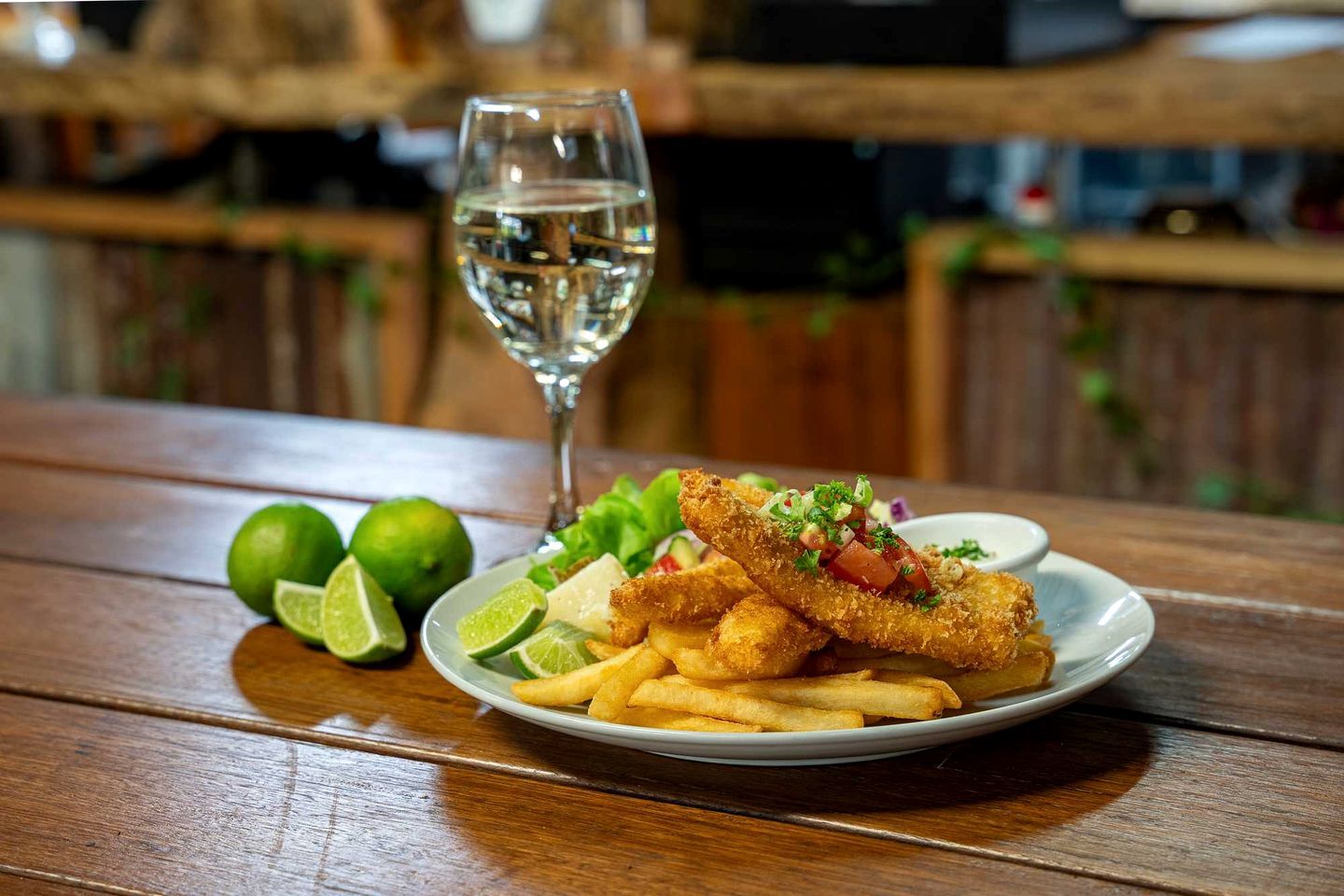 A plate of fried fish and fries with a glass of water, lime halves, and a small bowl of salad on a wooden table.