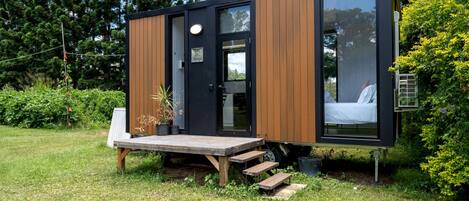 Une cabane moderne et compacte, avec une façade en bois, un lit visible à l’intérieur et un petit espace extérieur aménagé pour s’asseoir.
