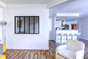 A modern living room with a large window, a white sofa, and a kitchen area in the background. - Chic 1BR by Louvre & Palais Royal (Paris)