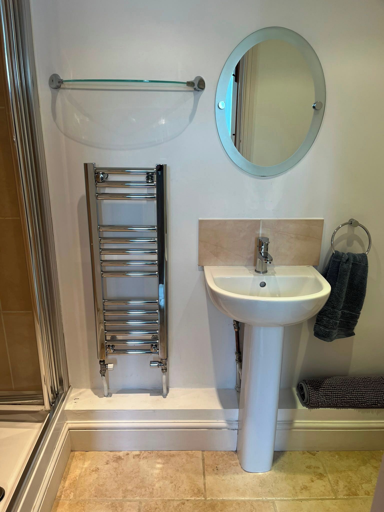 Une salle de bain avec une douche à parois de verre, un porte-serviettes en chrome, un miroir rond et un lavabo sur pied.