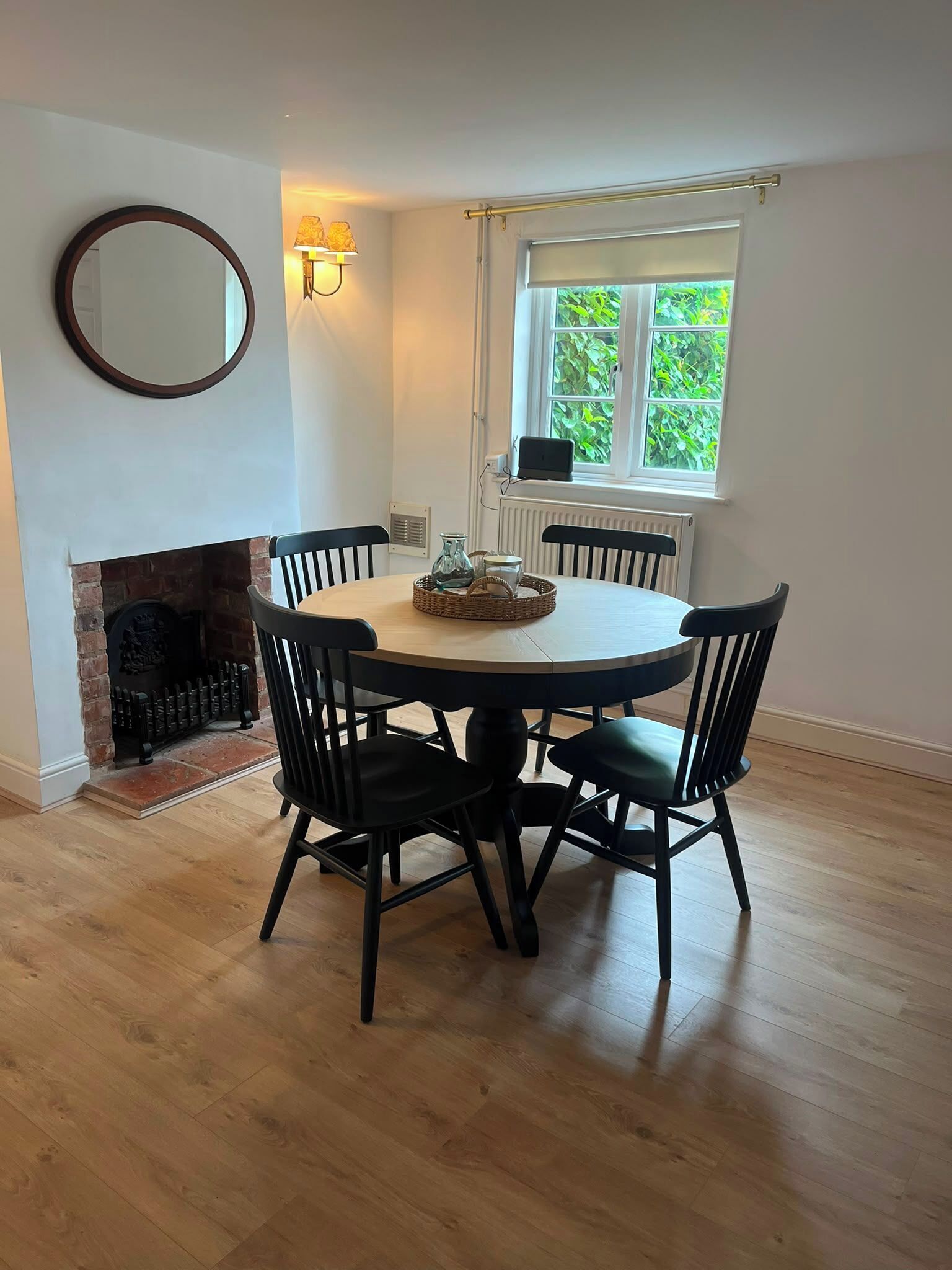 A dining area with a round table, four chairs, a fireplace, a window with a view of greenery, and wall-mounted lamps.