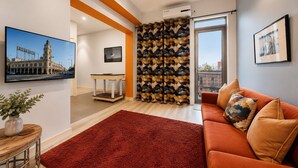 Una sala de estar moderna con alfombra roja, detalles en naranja, un sofá con cojines estampados, un televisor y vista a un paisaje urbano.